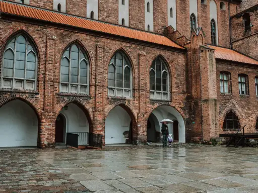 Zamek w Kwidzynie, z ceglaną fasadą i arkadowymi przejściami, w deszczowy dzień. Dwie postacie z parasolem spacerują po mokrym dziedzińcu.