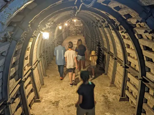 Rodzina zwiedza podziemia Sandomierz, idąc przez oświetlony tunel z kamiennymi ścianami i metalowymi wzmocnieniami.