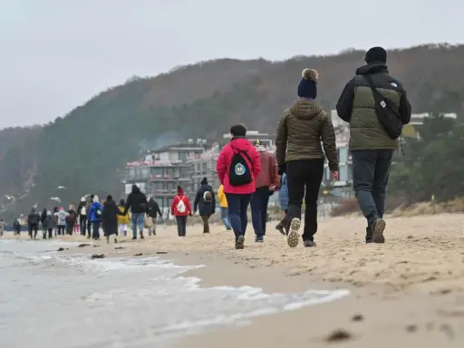 Spacer po plaży w chłodny dzień, idealny na długie weekendy 2026. Grupa ludzi w kurtkach i czapkach idzie wzdłuż morza.