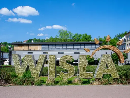 Wielkie litery "WISŁA" na rynku, ozdobione błyszczącymi elementami, na tle zieleni i budynków.