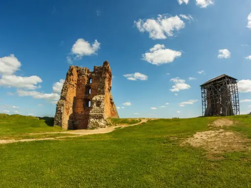 Ruiny ceglanego, opuszczonego zamku na zielonym wzgórzu pod błękitnym niebem z chmurami. Obok wieża w rusztowaniach.