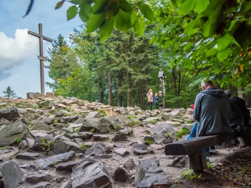 Kamienista ścieżka w **Górach Świętokrzyskich** prowadzi do drewnianego krzyża. Ludzie odpoczywają na ławkach wśród drzew.
