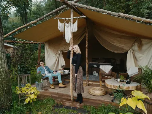 Para młoda relaksuje się w luksusowym kimura glamping namiocie. Mężczyzna siedzi na krześle, kobieta stoi przy drewnianym słupie.