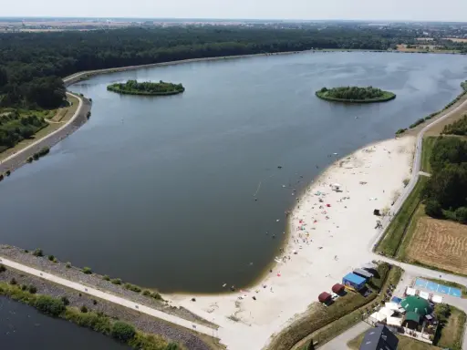 Letnie słońce nad jeziorem opolskim. Ludzie odpoczywają na piaszczystej plaży, pływają w wodzie. W oddali las i zabudowania.
