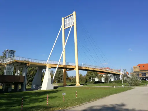 Żółty most pieszy, molo w Giżycku, z charakterystycznymi pylonami i linami, prowadzi do budynków nad jeziorem.