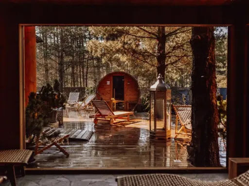 Drewniany domek w lesie, sauna beczka, leżaki i latarnia na mokrym tarasie.