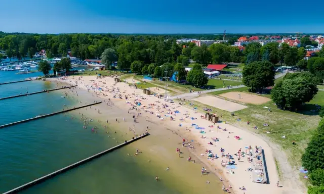 Słoneczna plaża w Giżycku tętni życiem. Ludzie odpoczywają na piasku, kąpią się w jeziorze i spacerują wzdłuż drewnianych pomostów.