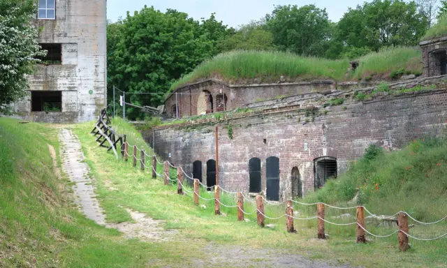 Fort Zachodni w Świnoujściu, zarośnięty trawą i otoczony zielenią, z widocznym starym budynkiem i historyczną artylerią.