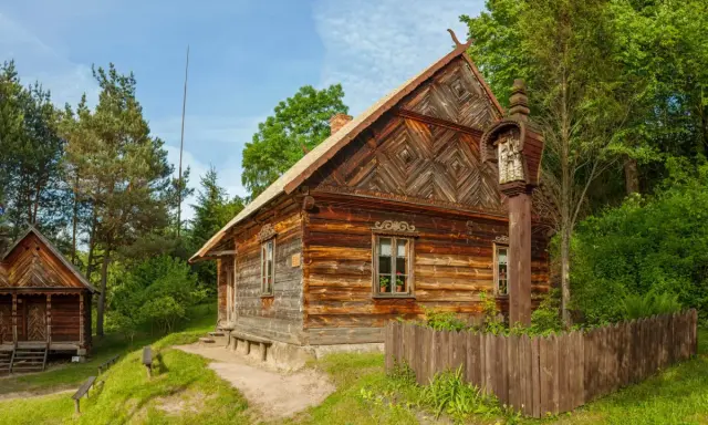 Drewniane chaty i kapliczka na słupie w jednym z **skansenów w Polsce**. W tle las i błękitne niebo.