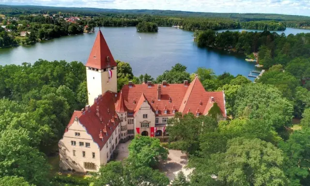 Zamek w Lubniewicach otoczony drzewami, nad jeziorem.