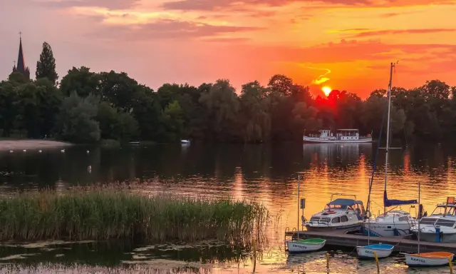 Zachód słońca nad jeziorem w Wielkopolsce. Łodzie przycumowane do pomostu, na wodzie pływają łabędzie, a w oddali widać statek wycieczkowy.