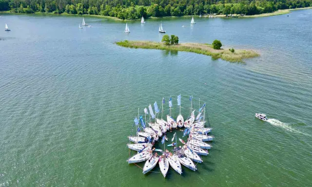 Żaglówki tworzą okrąg na jeziorze, idealne atrakcje Warmia i Mazury. W tle las i mała wyspa.