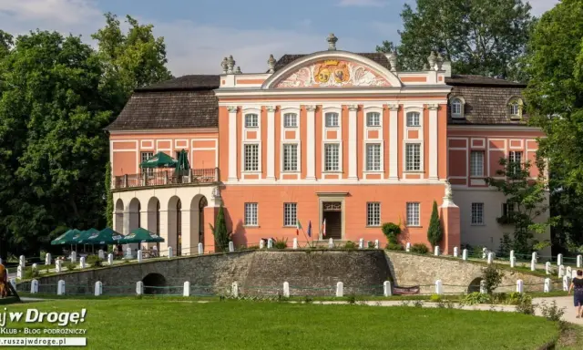 Kurozwęki pałac, z pomarańczową fasadą i białymi zdobieniami, otoczony zielenią. Na pierwszym planie mostek i trawnik.