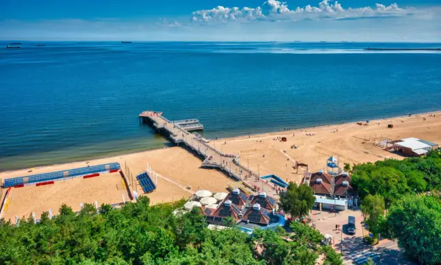 Idealne miejsce, gdzie na wakacje w sierpniu: piaszczysta plaża, molo, morze i zielone drzewa.