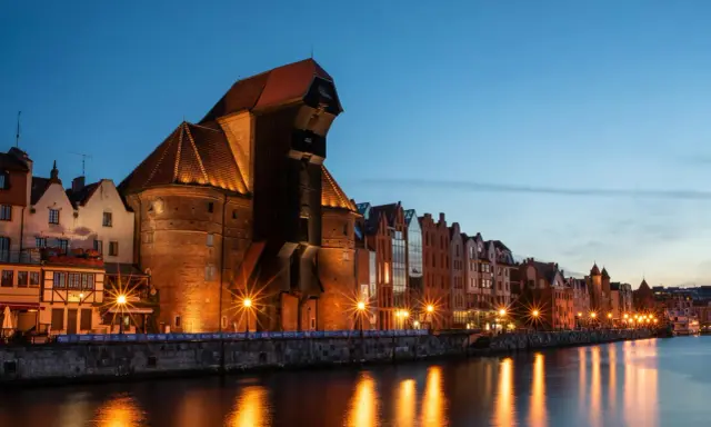 Gdańsk nocą. Historyczny Żuraw i kamienice nad Motławą, rozświetlone latarniami, odbijają się w wodzie.