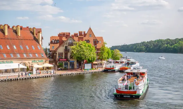 Port w Giżycku, gdzie cumują statki wycieczkowe, to jedna z głównych atrakcji miasta. W tle malownicze kamieniczki i zielone lasy.