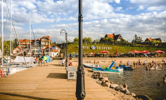 Słoneczna plaża nad jeziorem Śniardwy tętni życiem. Ludzie odpoczywają na piasku, kąpią się w wodzie, a obok cumują żaglówki.