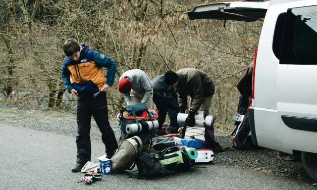 Grupa osób przygotowuje sprzęt do wyjazdu w góry. Plecaki, karimaty i torby czekają obok otwartego bagażnika.