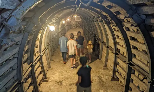Rodzina zwiedza podziemia Sandomierz, idąc przez oświetlony tunel z kamiennymi ścianami i metalowymi wzmocnieniami.