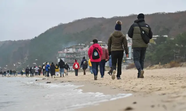 Spacer po plaży w chłodny dzień, idealny na długie weekendy 2026. Grupa ludzi w kurtkach i czapkach idzie wzdłuż morza.