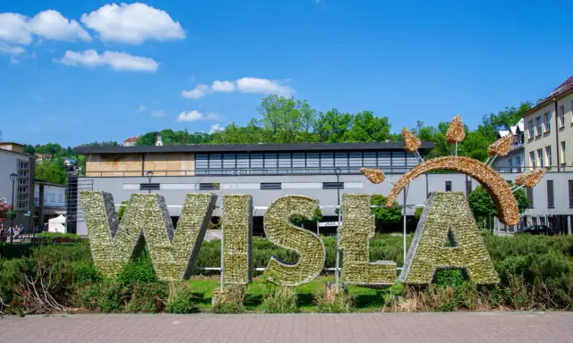 Wielkie litery "WISŁA" na rynku, ozdobione błyszczącymi elementami, na tle zieleni i budynków.