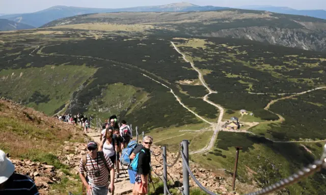 Grupa turystów wędruje po kamienistym szlaku w Karkonoszach. W oddali widać górzysty krajobraz i schronisko.