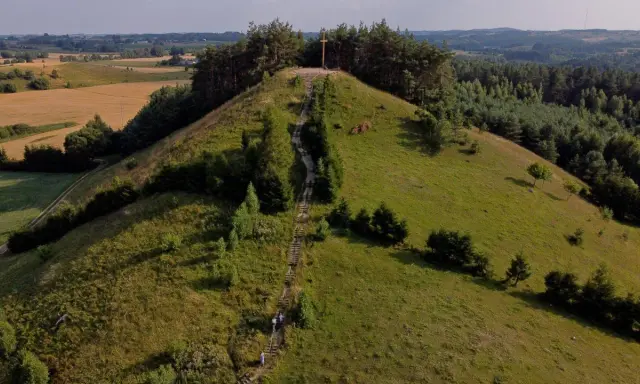 Wzgórze porośnięte trawą i drzewami, ze schodami prowadzącymi na szczyt, gdzie stoi drewniany krzyż. Widok na pola i lasy.