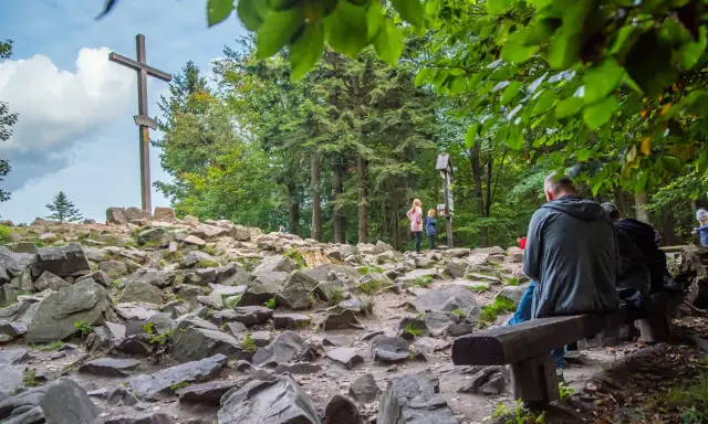 Kamienista ścieżka w **Górach Świętokrzyskich** prowadzi do drewnianego krzyża. Ludzie odpoczywają na ławkach wśród drzew.
