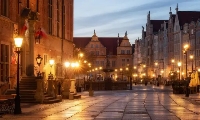 Wieczorny rynek Gdańsk oświetlony latarniami, z fontanną i zabytkowymi kamienicami.