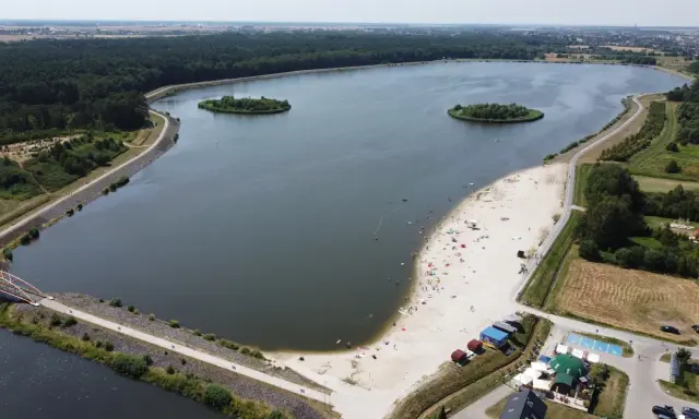Letnie słońce nad jeziorem opolskim. Ludzie odpoczywają na piaszczystej plaży, pływają w wodzie. W oddali las i zabudowania.