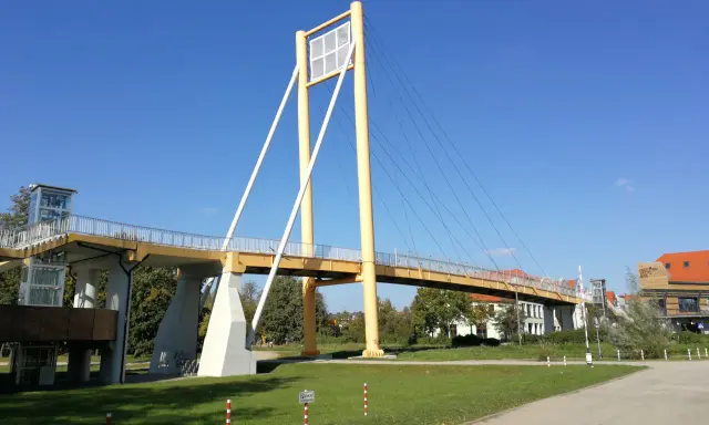 Żółty most pieszy, molo w Giżycku, z charakterystycznymi pylonami i linami, prowadzi do budynków nad jeziorem.