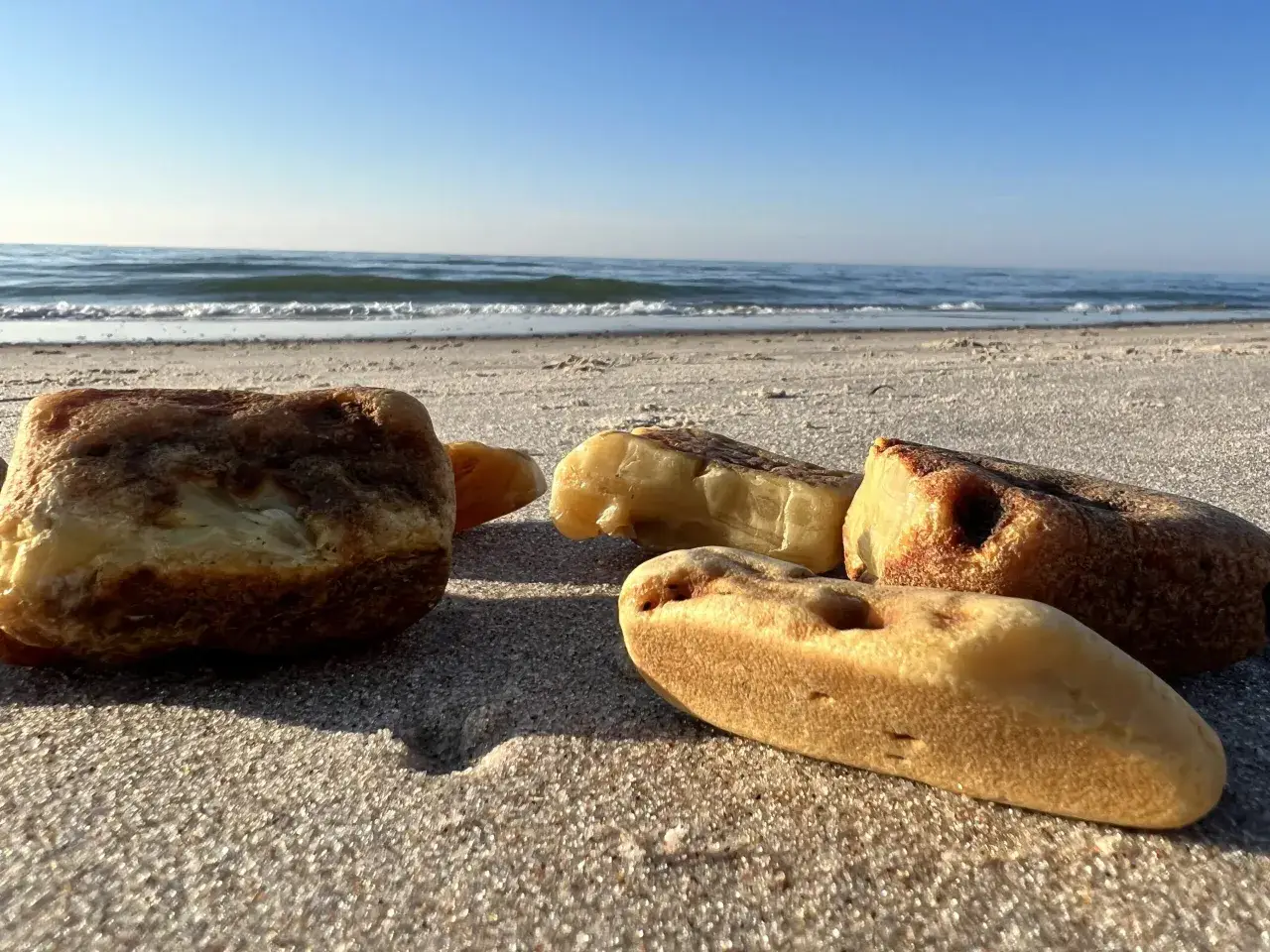Kilka kawałków bursztynu leży na piaszczystej plaży, w tle widać spokojne morze i błękitne niebo.