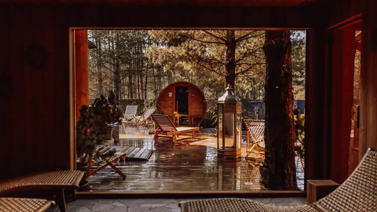 Drewniany domek w lesie, sauna beczka, leżaki i latarnia na mokrym tarasie.