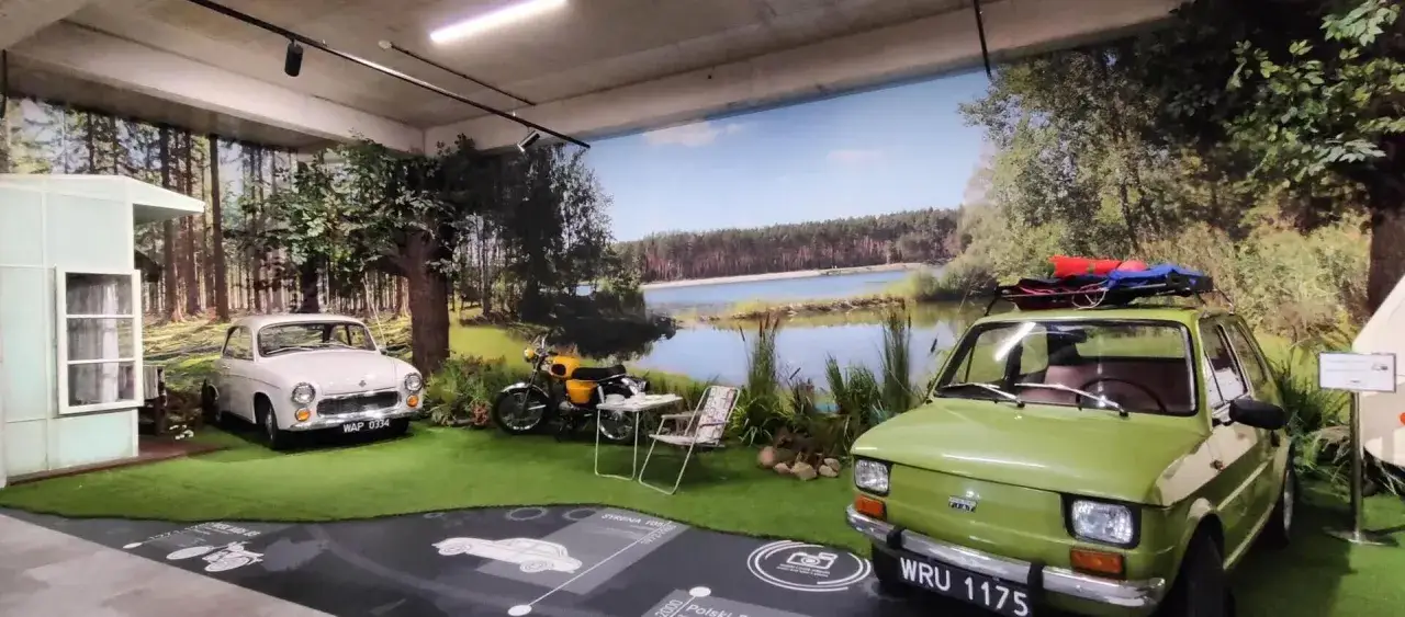 Wystawa w Muzeum Motoryzacji Śląsk: biały Syrena, żółty motorower i zielony Fiat 126p na tle leśnego krajobrazu z jeziorem.