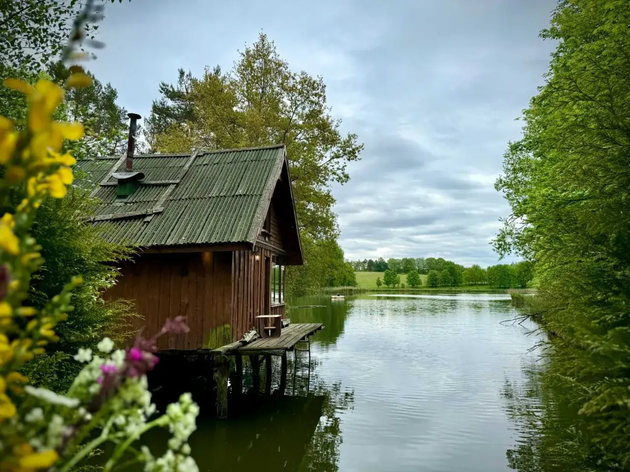 Ustronny domek nad jeziorem, z drewnianym pomostem i kominem. Wokół bujna zieleń i spokojna tafla wody.