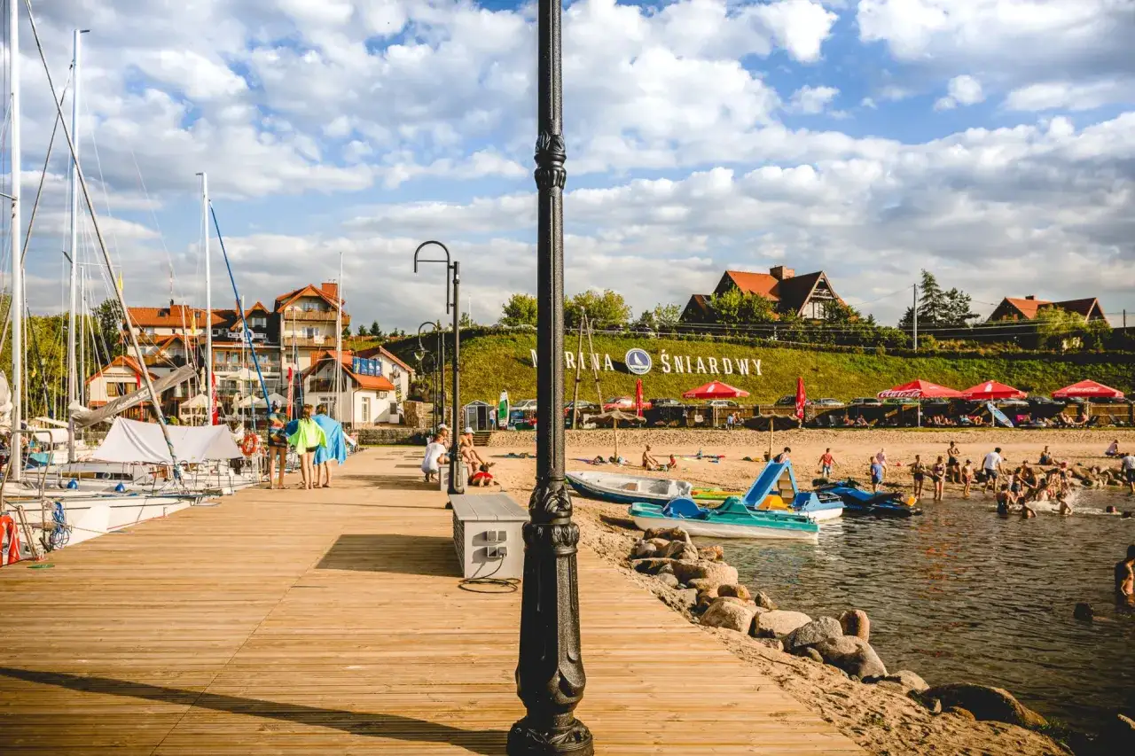 Słoneczna plaża nad jeziorem Śniardwy tętni życiem. Ludzie odpoczywają na piasku, kąpią się w wodzie i pływają łódkami.