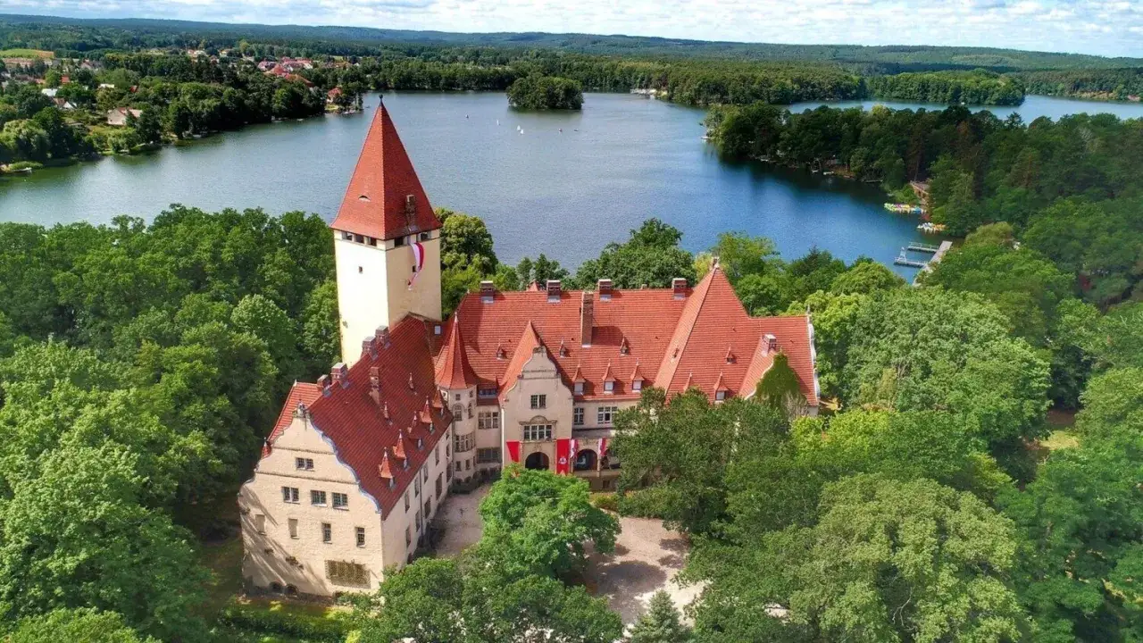 Zamek w Lubniewicach, otoczony zielenią, z czerwonymi dachami, góruje nad jeziorem.