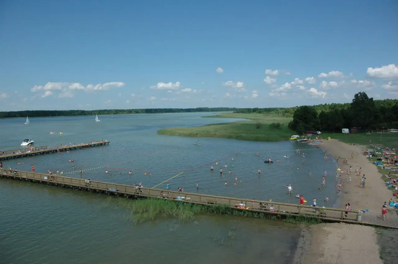 Letnie popołudnie nad jeziorem Roś. Ludzie kąpią się, opalają na plaży i spacerują po drewnianych pomostach.