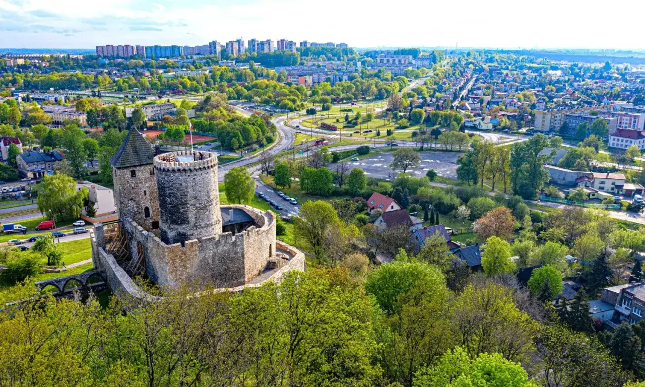 Kamienny zamek góruje nad miastem, przypominając o historii zamków na Śląsku. W tle rozciąga się zielona panorama z budynkami i drogami.