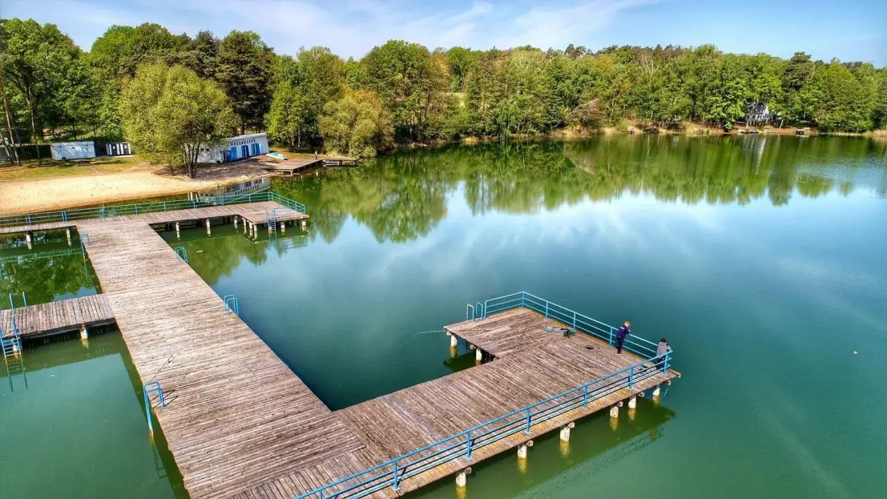 Drewniane pomosty nad spokojnymi jeziora zielona g&oacute;ra, otoczone zielenią. Dwa wędkarze łowią ryby.