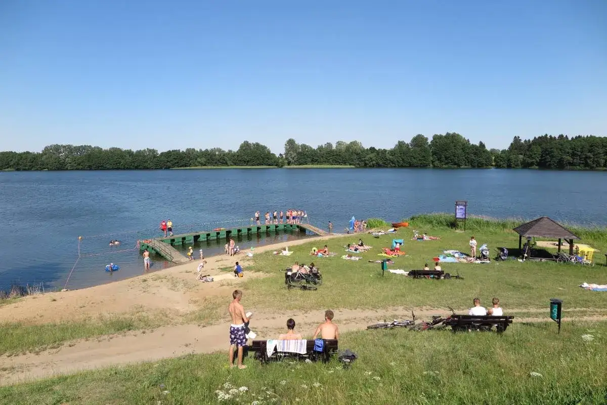 Ludzie odpoczywają nad wodą, niektórzy kąpią się, inni opalają na trawie. Jezioro Drawsko w słoneczny dzień.