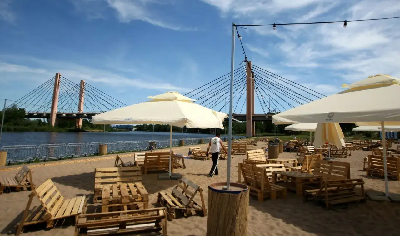 Plażowy bar z drewnianymi meblami i parasolami, w tle most. Idealne trójmiasto atrakcje dla dorosłych szukających relaksu nad wodą.