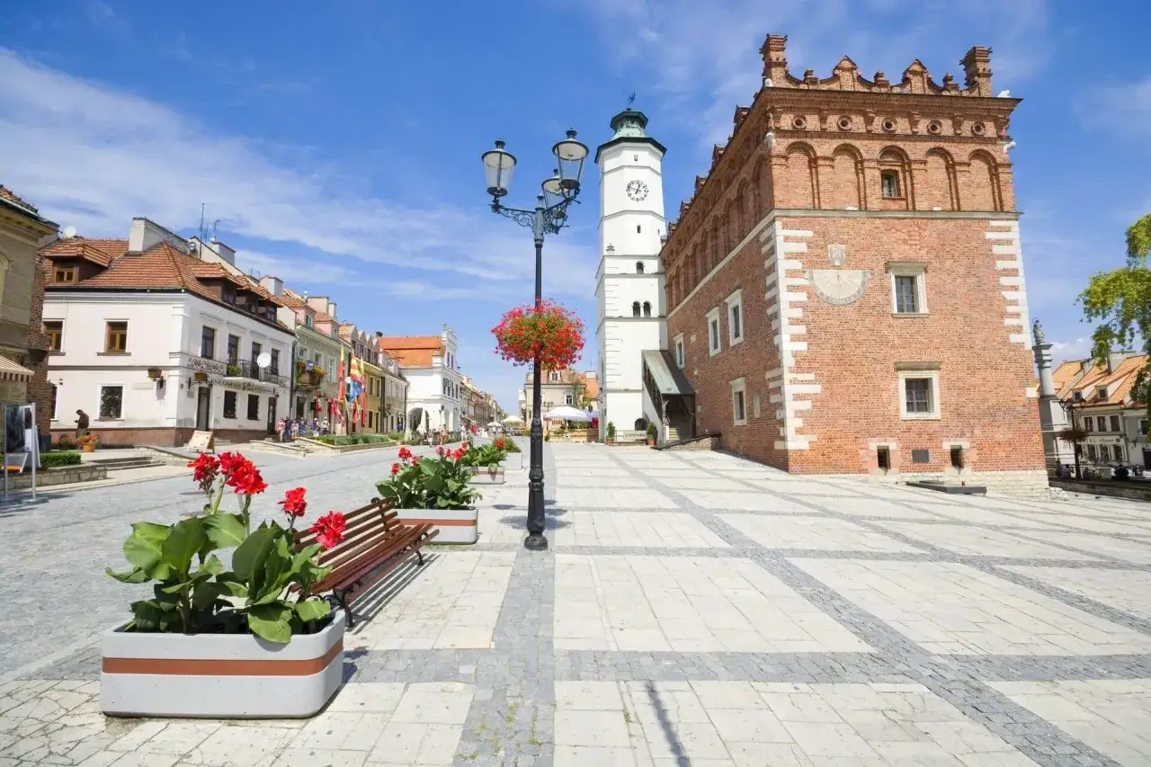 Słoneczny dzień na najładniejszym rynku w Polsce. Ceglany ratusz, biała wieża zegarowa i kolorowe kamienice tworzą malowniczy widok.