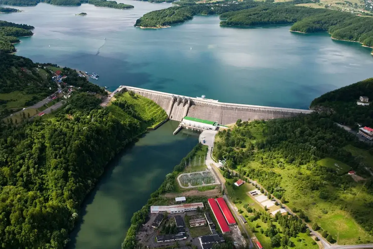Widok z lotu ptaka na zaporę i elektrownię Solina. Woda w jeziorze jest turkusowa, otoczona zielonymi wzgórzami.