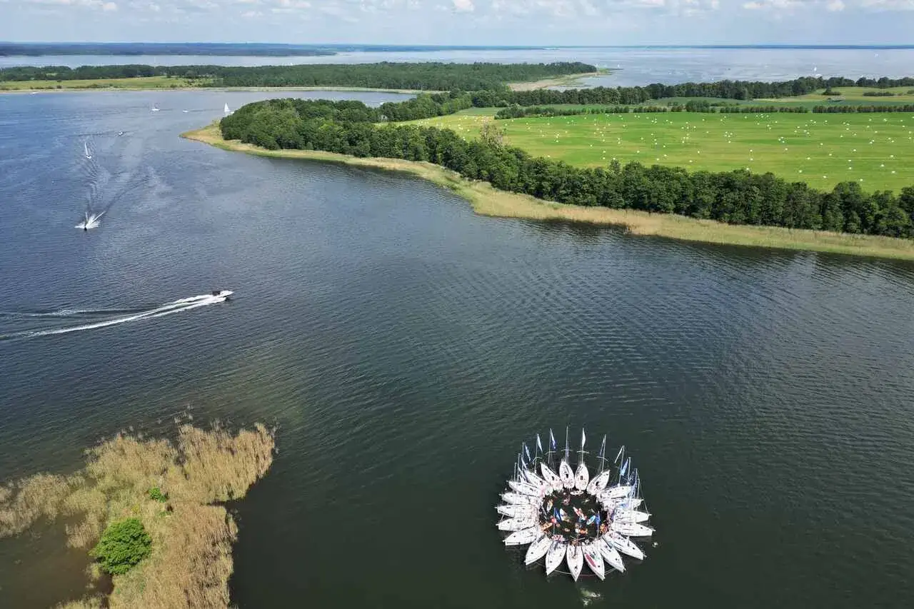 Mazury słyną z jezior i żeglarstwa. Na wodzie formacja łodzi przypomina kwiat, a w oddali widać pola i lasy.