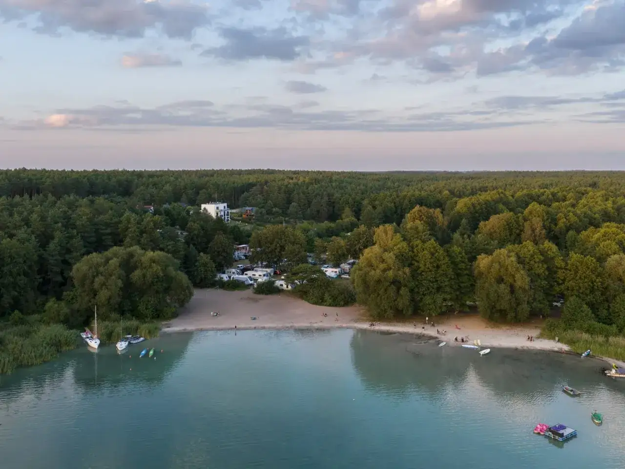 Widok na piaszczystą plażę i turkusową wodę, otoczoną lasem. W oddali widać kemping z kamperami. To miejsce to prawdopodobnie najczystsze jezioro w Polsce.