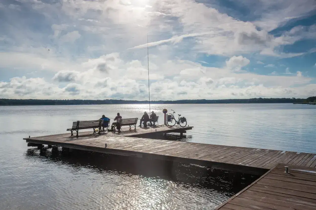 Ludzie łowią ryby na pomoście nad jeziorem Sławskim. Słońce przebija się przez chmury, a na brzegu stoi rower.