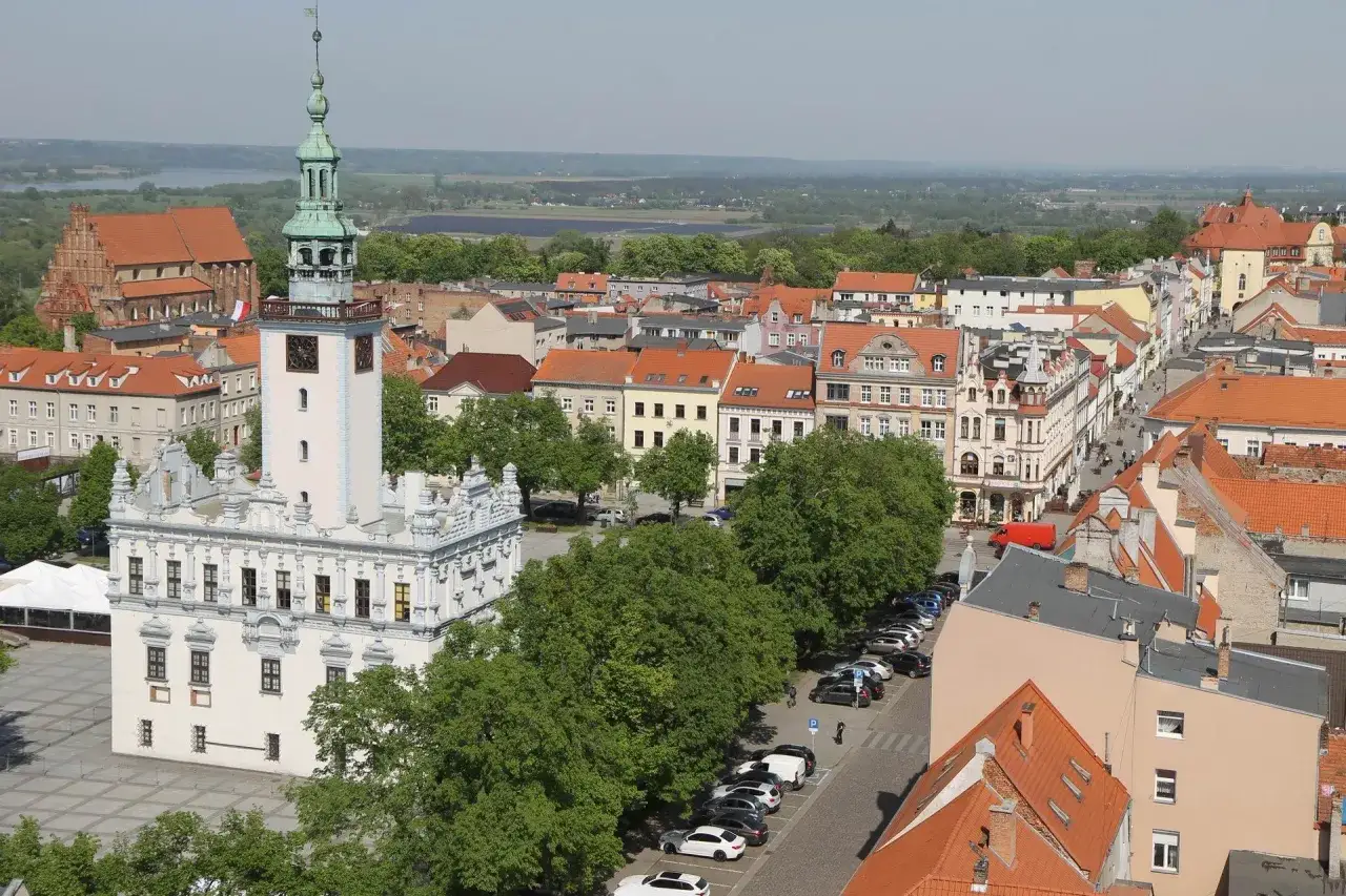 Widok na rynek w Chełmnie, z ratuszem i wieżą zegarową. W tle zabudowa miejska i zieleń. Trasa spacerowa przez miasto.