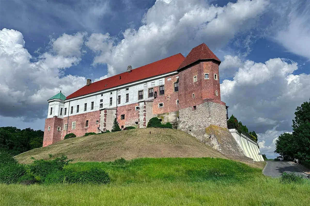 Zabytkowe miasto nad Wisłą, zamek na wzgórzu, z czerwonym dachem i ceglanymi murami, pod błękitnym, chmurnym niebem.
