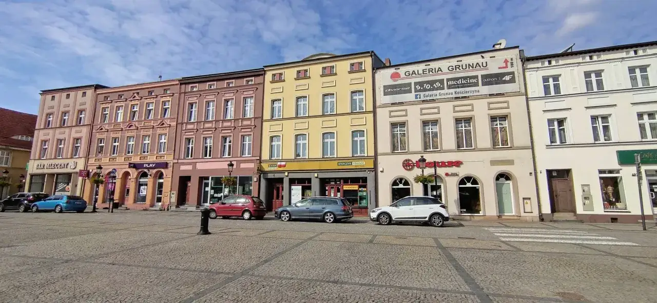 Kolorowe kamienice na kościerskim rynku, z samochodami i przejściem dla pieszych.